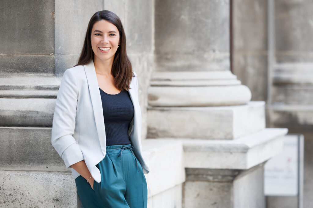 Auf dem Bild ist Rahel More. Eine lächelnde junge Frau mit langen Haaren und weißem Blazer. Sie steht angelehnt an eine Saule. 